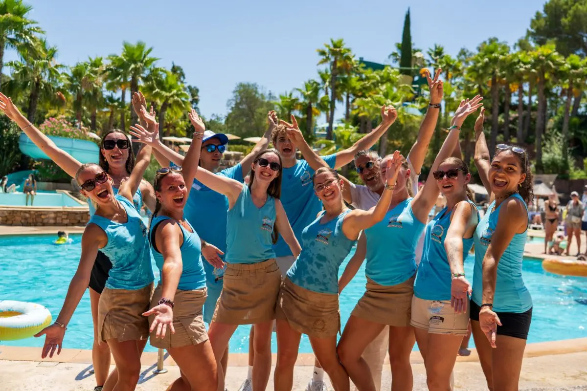The Esterel Caravaning team, arms raised and all smiles, by the pool beneath the palm trees